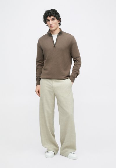 Pull en tricot marron avec un col zippé demi-long, associé à un pantalon large gris clair et des baskets blanches. Le tissu semble doux et texturé.