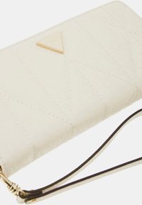 Valkoinen tilkkupussukka, jossa on texturoitu pinta, varustettuna kullanvärisellä kolmiomaisella Guess-logolla ja irrotettavalla rannehihnalla. Kullanväriset varusteleikkerit ovat näkyvissä.