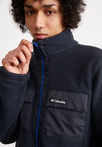 Black fleece jacket with blue zipper and patches. Features a high collar and front pocket. Soft texture, warm material, casual design.