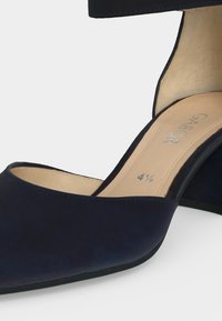 Gros plan d’une chaussure à talon haut bleu marine avec bride à la cheville, semelle intérieure beige et taille 4½ marquée à l’intérieur.