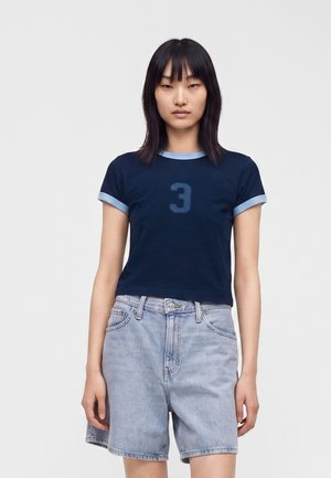 Mujer con cabello negro liso lleva una camiseta azul marino con el número 3 y shorts vaqueros azul claro sobre un fondo liso.