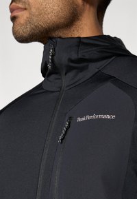Czarna sportowa kurtka z wysokim kołnierzem, suwakiem na kieszeni na piersi oraz logo "Peak Performance". Gładki materiał o dopasowanym kroju.