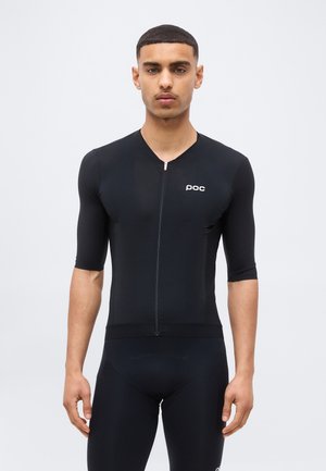 Junger Mann steht frontal und trägt ein eng anliegendes schwarzes Radtrikot und Shorts mit "poc"-Logo auf der Brust.