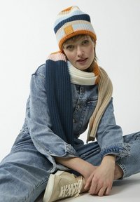 Gebreide beanie met meerkleurige strepen en een gestructureerde sjaal. Beide hebben oranje, marineblauwe en crèmekleurige tinten. Casual spijkeren outfit en beige sneakers.