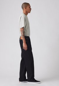 Tricou gri cu mânecă scurtă, asortat cu blugi negri, drepți. Detalii notabile includ o etichetă din piele crem pe blugi și adidași negri.