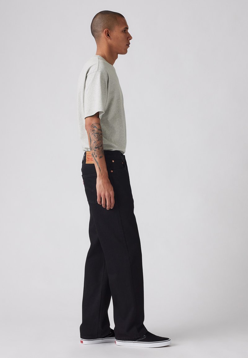 T-shirt gris à manches courtes associé à un jean noir ample. Les détails notables incluent un patch en cuir beige sur le jean et des baskets noires.