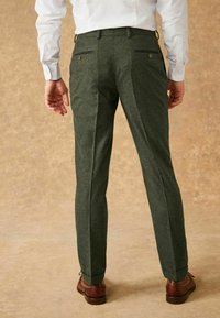 Pantalon texturé vert avec une coupe ajustée, comportant des poches arrière avec fermetures éclair et accents en bouton, associé à des chaussures habillées marron.