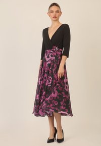 Mustat mekko syvällä kaula-aukolla ja kolmannen neljänneksen hihoilla, jossa on kukkakuosiset violetti hame ja kangaskukka-akcentti vyötäröllä.