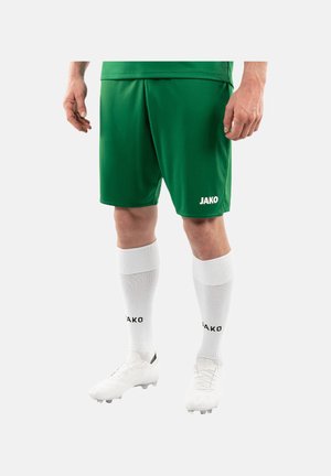 Mandlig atlet iført grønne fodboldshorts, hvide knæstrømper og hvide fodboldstøvler, stående med hænder afslappet langs siden.