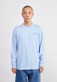 Camisa de manga longa azul claro, feita de algodão, com um pequeno logótipo roxo "nike" no lado esquerdo do peito e um corte relaxado. O fundo é neutro.