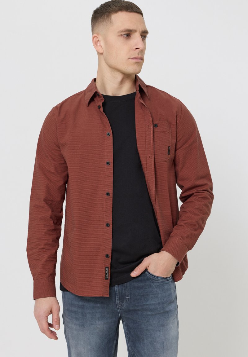 Homme portant une chemise boutonnée couleur rouille sur un t-shirt noir, un jean bleu, debout avec une main dans la poche, regardant sur le côté.