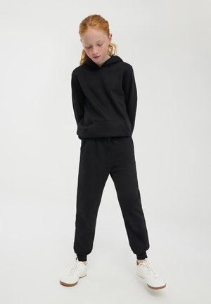 Completo con felpa nera e pantaloni della tuta realizzati in tessuto morbido, dotato di tasca frontale, polsini elasticizzati e vita con cordoncino. Le sneakers bianche completano l'outfit.