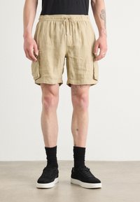 Pantaloni corti in lino beige con elastico in vita e cordoncino, dotati di due tasche laterali e una vestibilità comoda. Indossati con calze e scarpe nere.