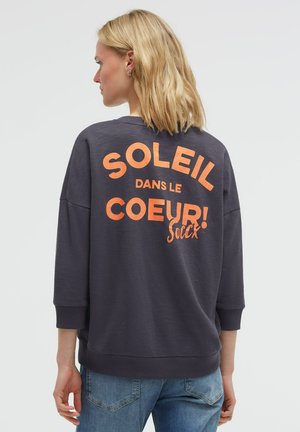 Blonde woman wearing a dark gray sweatshirt with orange text "SOLEIL DANS LE COEUR! Soccx" on the back, paired with blue jeans.