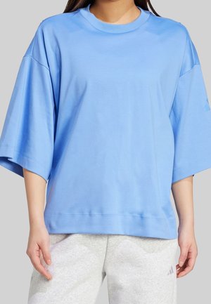 Personne portant une chemise ample à manches courtes bleu clair et un pantalon de survêtement gris, debout devant un fond uni.
