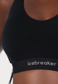 Black sports bra made of soft material, featuring a rounded neckline and a wide band with "icebreaker" logo in gray. Smooth texture.