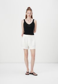 Canotta nera a coste con spalline larghe, abbinata a pantaloni corti di lino bianchi e ciabatte nere. Design semplice, outfit estivo casual.