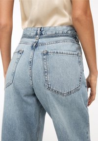 Jeans de denim de talle alto en color azul claro con bolsillos traseros, utilizados por una persona con una blusa sin mangas en color crema, de pie contra un fondo blanco.