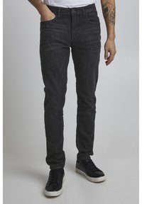 Dark denim jeans with a slim fit, featuring five pockets and a zip-fly closure. Worn with black sneakers. Textured fabric detail visible.