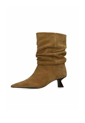 Bruine suède laarzen tot halverwege de kuit, slordige stijl, met een spitse neus en lage, schuine zwarte hak tegen een witte achtergrond.