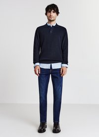 Marineblauer Strickpullover über einem hellblauen Kragenhemd, kombiniert mit dunkelblauen Slim-Fit-Jeans und schwarzen Leder-Stiefeletten, stehend vor einem schlichten Hintergrund.