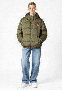 Olijfgroene pufferjas met een doorgestikt design, voorzien van een geborduurde "I ❤️ You" op de linkerborst, gecombineerd met losse blauwe jeans en witte sneakers.