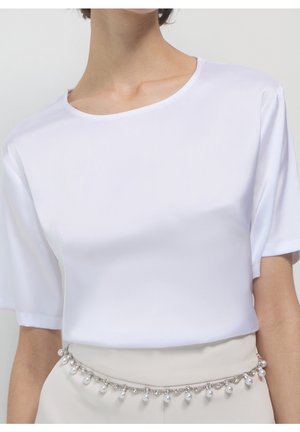 Femme portant un t-shirt blanc uni à manches courtes rentré dans un pantalon beige avec un accessoire chaîne de ceinture argentée et perles.