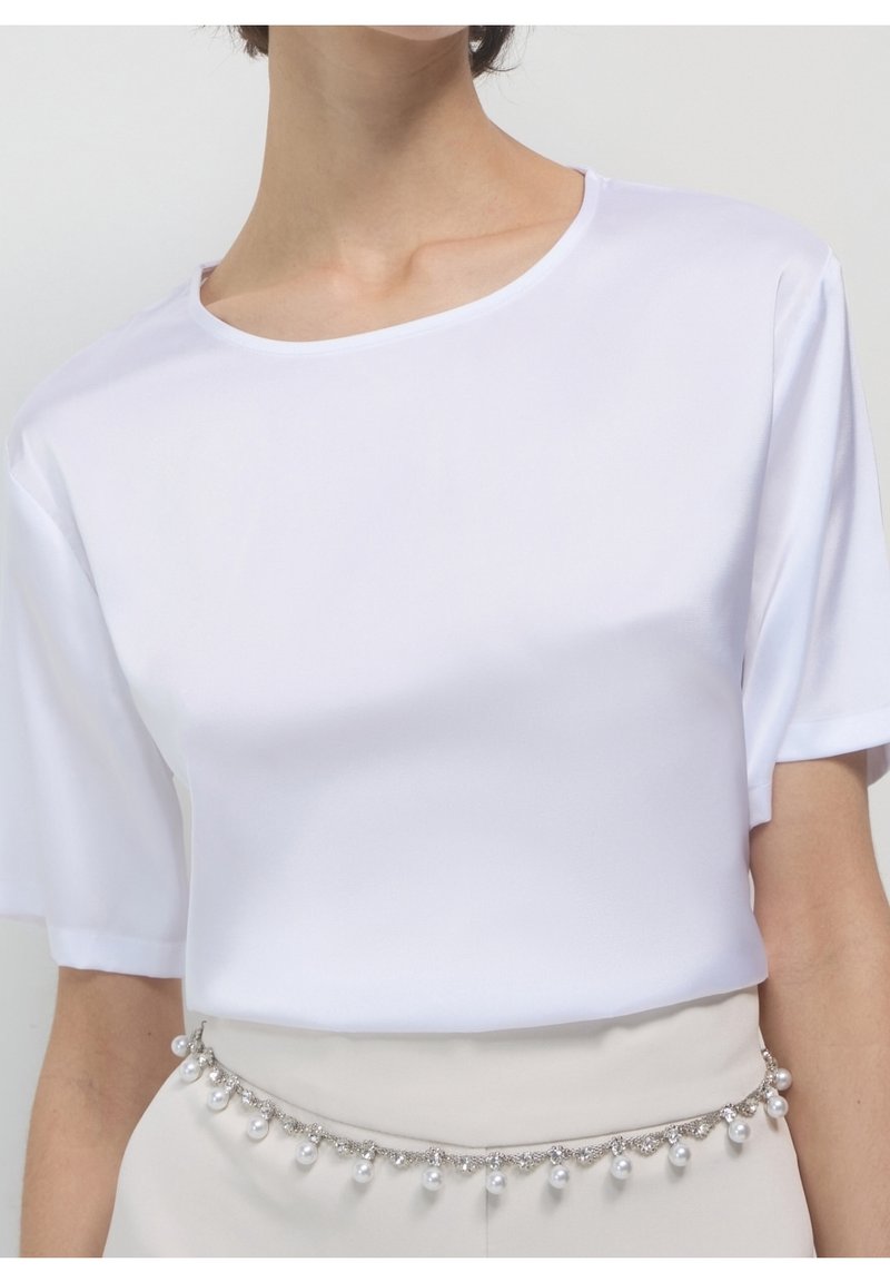 Femme portant un t-shirt blanc uni à manches courtes rentré dans un pantalon beige avec un accessoire chaîne de ceinture argentée et perles.