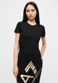 Μαύρο εφαρμοστό φόρεμα κοντομάνικο από απαλό βαμβάκι, με χρυσό λογότυπο γραφικού και διαγώνιο σχέδιο ρίγας στο μπροστινό μέρος. Κοντά μανίκια.