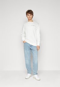 Hvid langærmet sweatshirt med teksten "GANT 1948", parret med lyseblå jeans og hvide sneakers, stående på en neutral baggrund.