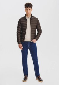 Brązowa kurtka typu puffer z poziomym pikowaniem, w połączeniu z beżowym polo i ciemnoniebieskimi dżinsami. Brązowe buty na sznurówki dopełniają stylizację.