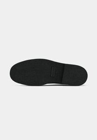 Black rubber shoe sole, textured surface, oval shape, rounded toe with a slight lift, featuring a brand logo and patterned grip.