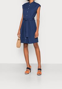 Navyblaues Hemdblusenkleid mit taillierter Gürtellung, kurzen Ärmeln und Knopfverschluss. Kombiniert mit orangefarbenen und navyblauen Riemchenschuhen. Geflochtene Tasche.