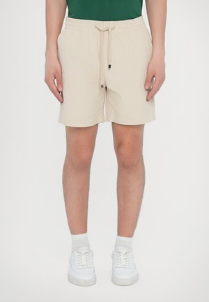 Beige bomullsshorts med elastisk midje og snøring. Har sidelommer og rett snitt, kombinert med hvite sneakers.