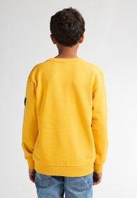Sudadera de algodón amarilla con puños y dobladillo acanalados, que presenta un pequeño parche de logo en la manga izquierda y un corte relajado. Jeans visibles por debajo.