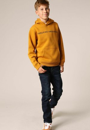 Gele hoodie met kap en zwarte tekst "DEELUXE ORIGINAL", voorzak, gecombineerd met donkerblauwe spijkerbroek en oranje sneakers.