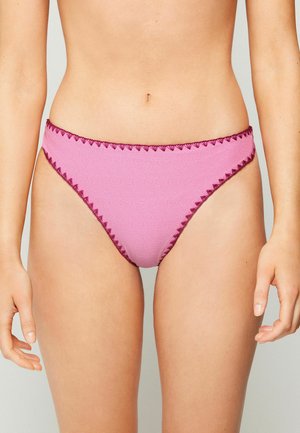 Frau trägt pinke Bikinihose mit hohem Beinausschnitt und dunkelrosa Zickzackbesatz, dargestellt von der Taille bis zur Mitte des Oberschenkels vor hellem Hintergrund.