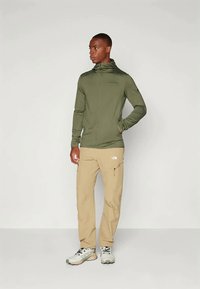 Άνδρας που στέκεται σε μπεζ cargo παντελόνι, ελαφριά υποδήματα πεζοπορίας και πράσινο μπουφάν με φερμουάρ και ψηλό γιακά, κοιτάζοντας πλάγια με το ένα χέρι στην τσέπη.