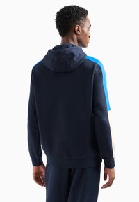 Sudadera con capucha azul marino con detalles en azul y blanco, que presenta un ajuste relajado, puños acanalados y una textura suave y lisa.