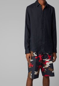 Muž oblečený v černé košili s dlouhým rukávem a šortkách do kolen s maskáčovým vzorem v červené, hnědé, bílé a černé barvě.