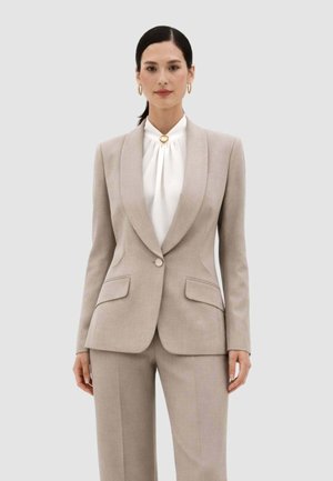 Donna con completo beige sartoriale composto da blazer monopetto e pantaloni abbinati, con blusa bianca a collo alto e orecchini a cerchio dorati.