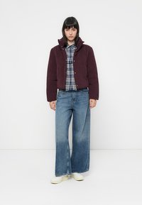 Bordeaux gewatteerde jas met een hoge kraag, gelaagd over een blauw geruit overhemd. Gecombineerd met een wijde blauwe spijkerbroek en witte sneakers.