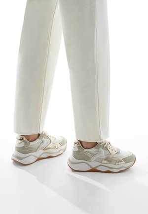 Sneakers chunky in un beige chiaro con tomaia in rete bianca, dettagli dorati e sovrapposizioni testurizzate, abbinati a pantaloni dritti bianchi.