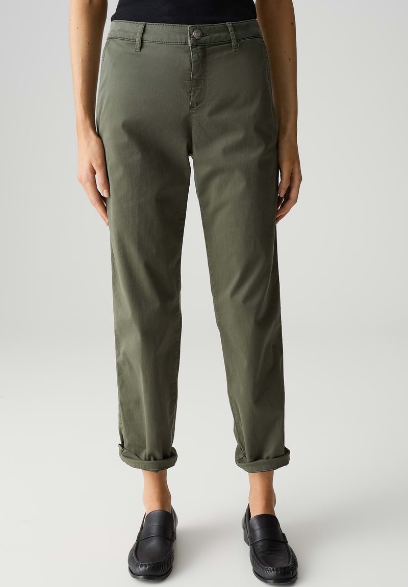 Pantaloni tapered in cotone misto verde oliva con orli arrotolati, chiusura anteriore con bottone e tasche laterali, abbinati a scarpe nere slip-on.