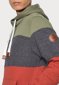 Sudadera con capucha con un diseño de bloques de color en verde, azul marino y óxido. Presenta cordones blancos y un parche de logo de cuero marrón en la manga.