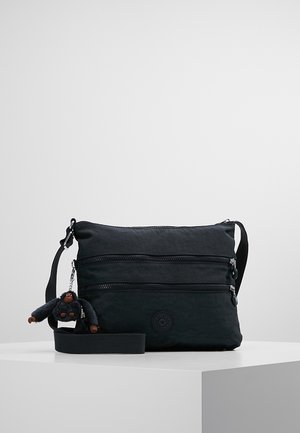 Czarna torebka na ramię z dwoma przednimi kieszeniami na suwak oraz małą pluszową zawieszką w kształcie małpki przymocowaną do paska, prezentowana na białej platformie.