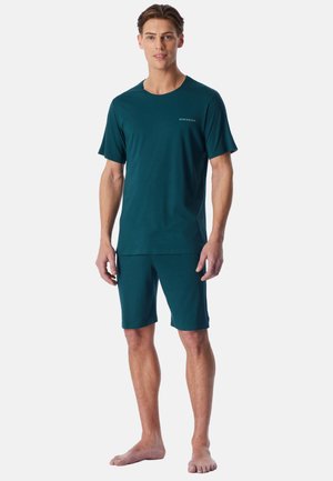 Homme debout pieds nus, portant une chemise à manches courtes bleu sarcelle foncé et un short assorti jusqu’aux genoux sur un fond uni.