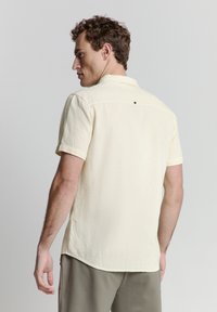 Crème-kleurig kortomshirt met een gestructureerde stof, colbertkraag en een knoopdetail aan de achterkant. Rechte zoom met licht afgeronde randen.