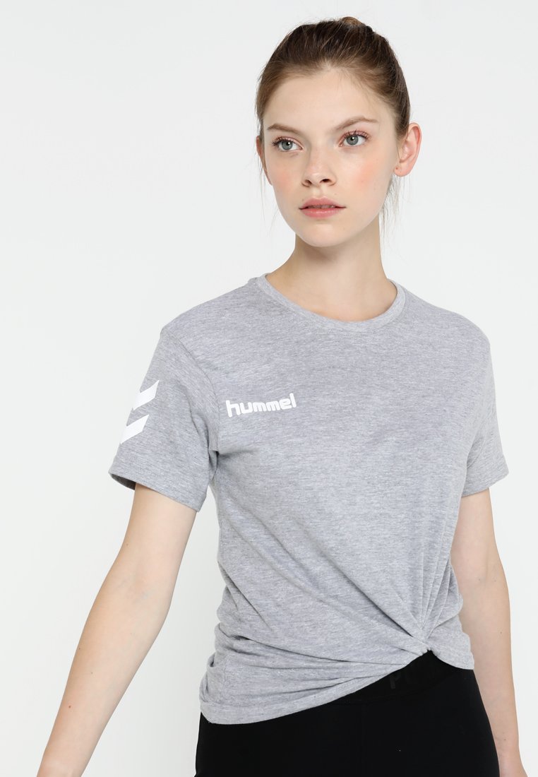 Junge Frau trägt ein hellgraues Hummel-T-Shirt mit seitlichem Knoten, steht vor einem einfarbigen weißen Hintergrund und schaut leicht nach rechts.