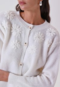 Cardigan bianco lavorato a maglia con applicazioni floreali testurizzate, scollo rotondo e bottoni, caratterizzato da una consistenza morbida e pelosa.